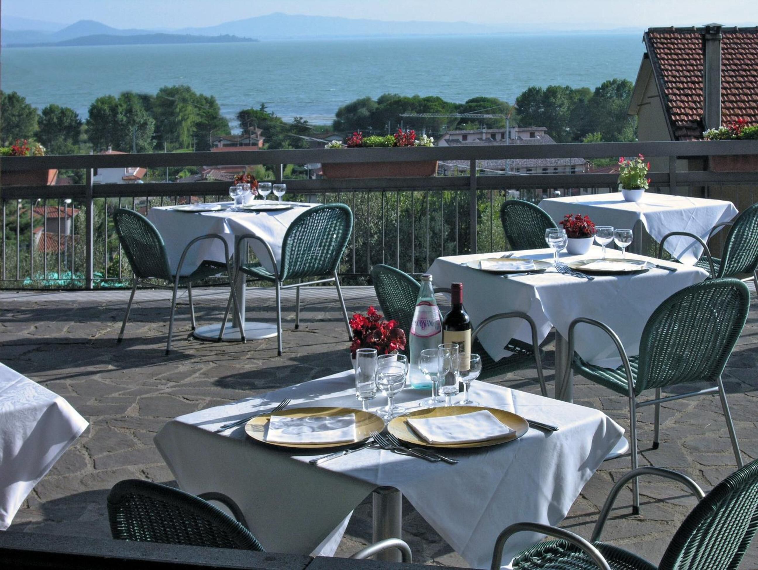 Des tables de jardin, recouvertes de nappes blanches, entourées de chaises vertes et agrémentées de verres à vin, sont disposées sur une terrasse offrant une vue sur la mer et les montagnes en arrière-plan.