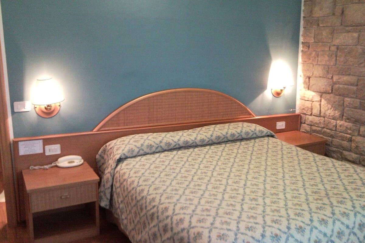 A bedroom with a wooden headboard, a patterned bedspread, two bedside lamps, a nightstand with a telephone, and a brick wall.