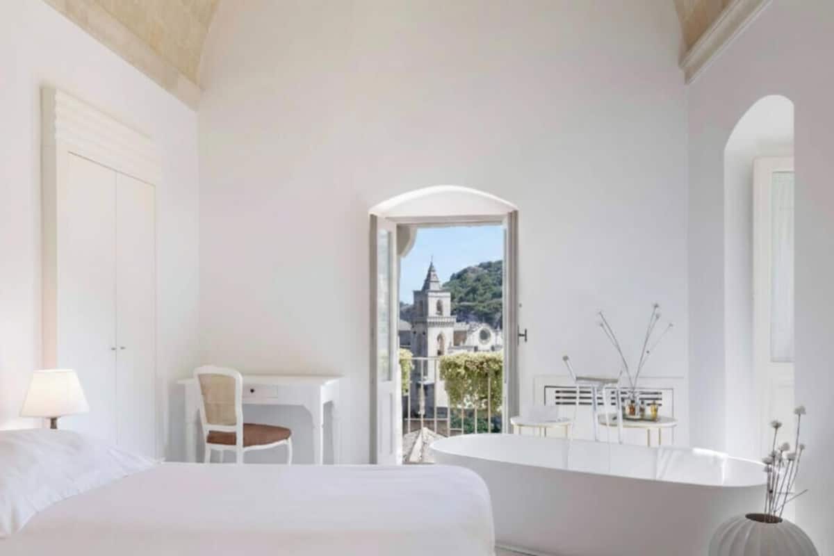 A hotel room with a bed, a desk, a chair, a bathroom with a bathtub, and a view of a church through an open door.