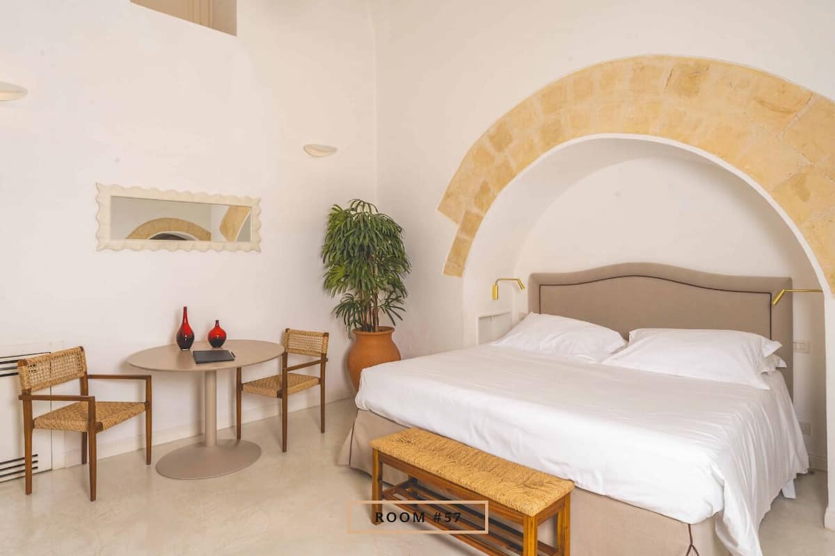 A bedroom with a bed, a round table with chairs, a potted plant, and a mirror on the wall.