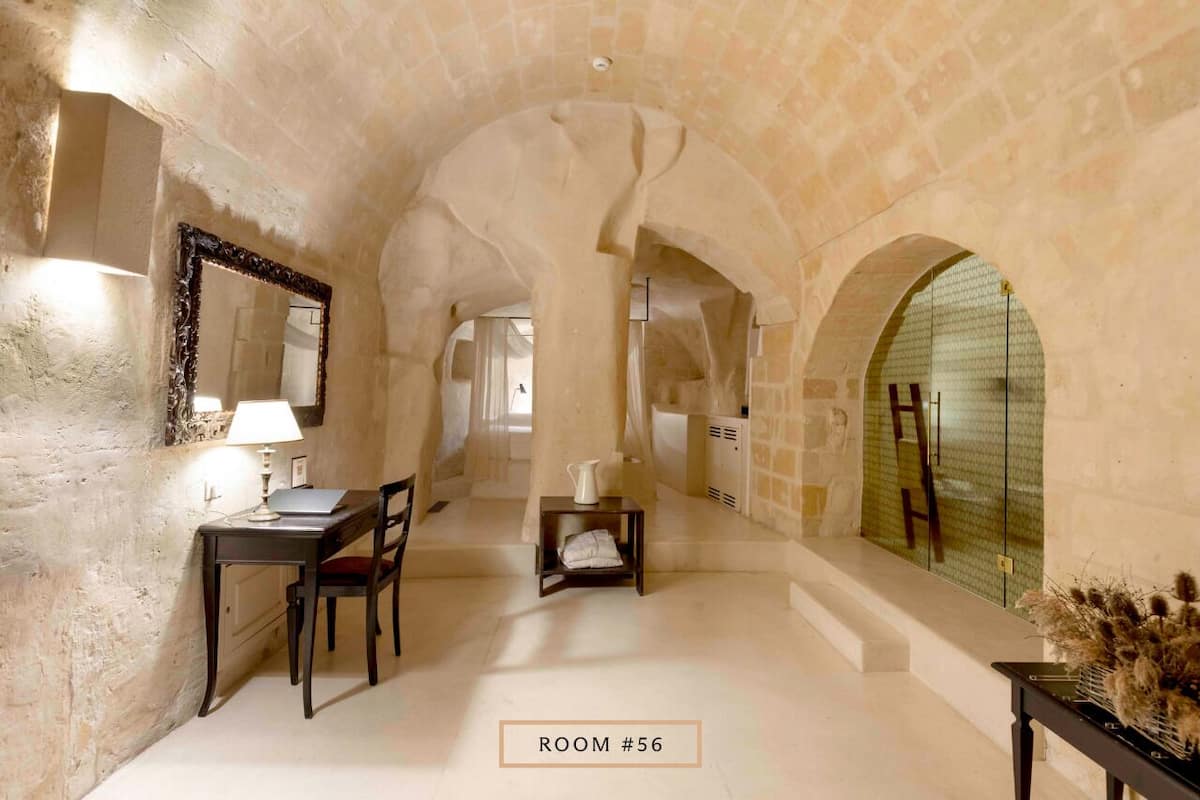 A room with a stone wall, a desk with a lamp, a chair, a small table with a pitcher, and a vase with dried flowers.