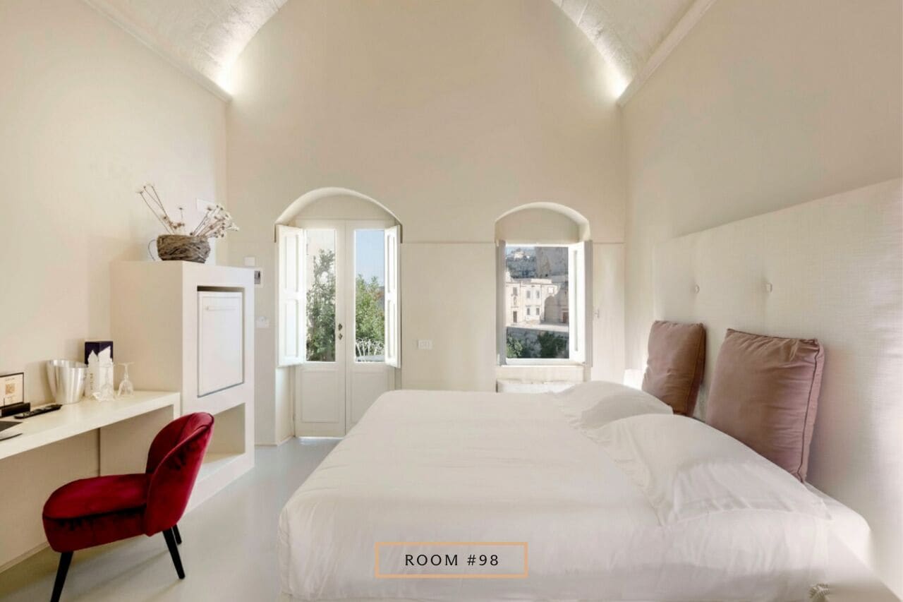 A bedroom with a large bed, a desk with a chair, and two windows with arched tops.