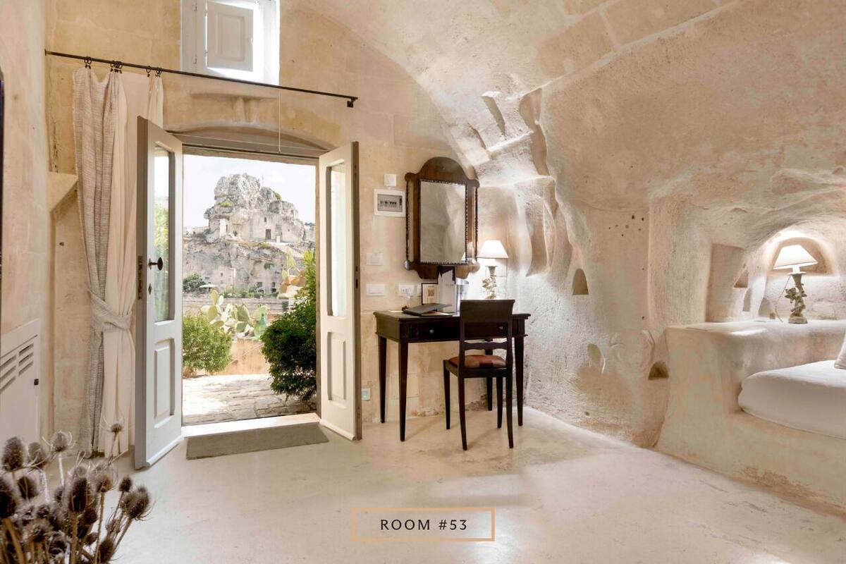 A room with a stone wall, a desk with a chair, a mirror, and a view of a mountainous landscape.