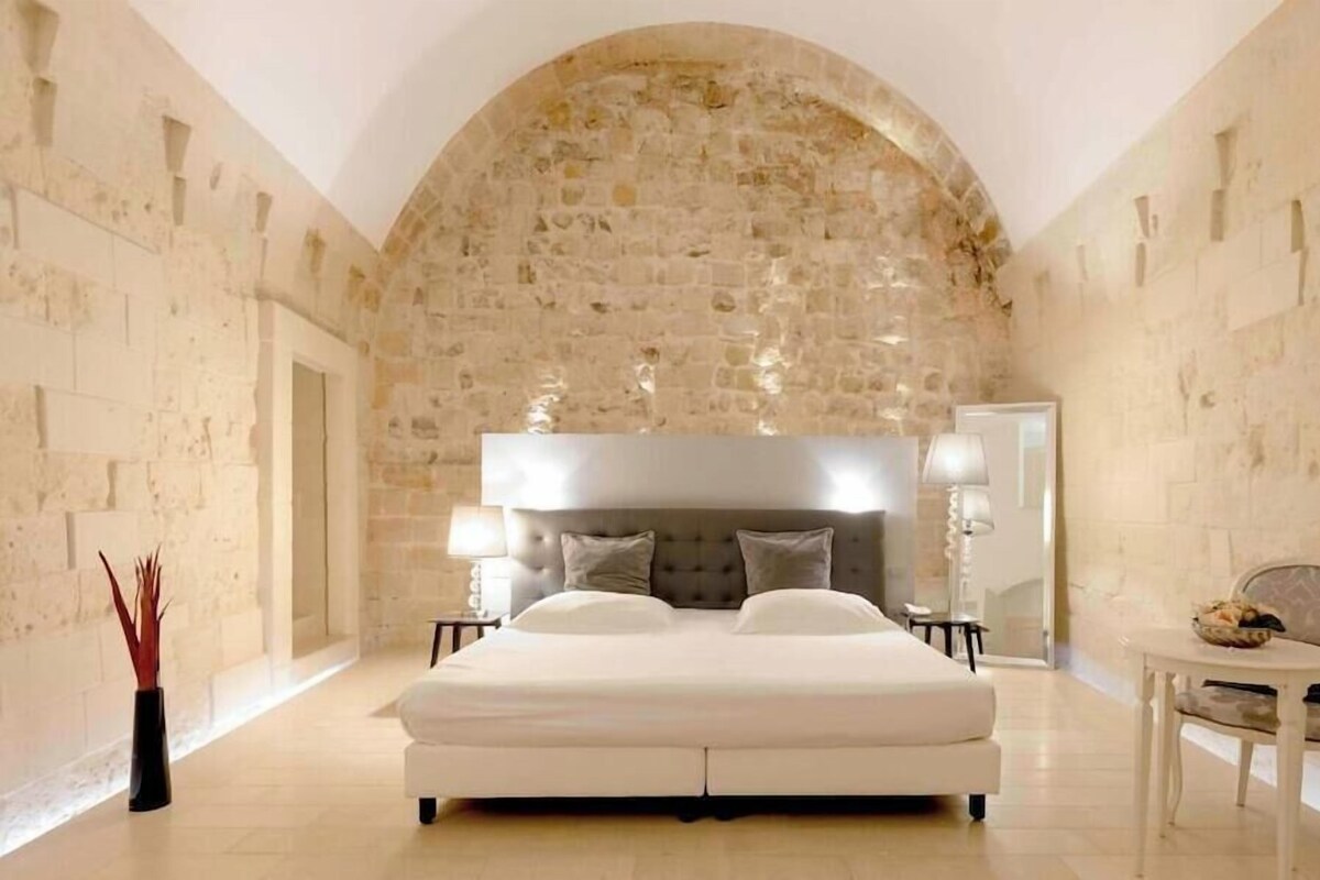 A bedroom with a stone arch, a large bed, bedside lamps, and a small table with a chair.