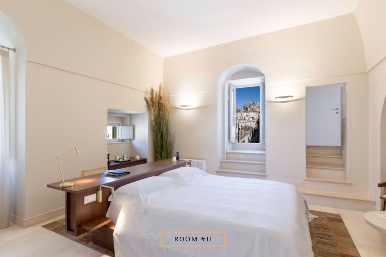 Une chambre d’hôtel moderne avec un grand lit, un bureau avec une lampe, un miroir et une vue sur le paysage urbain, visible à travers une fenêtre en arc de cercle.