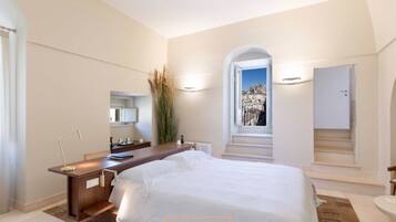 Une chambre d’hôtel moderne avec un grand lit, un bureau avec une lampe, un miroir et une vue sur le paysage urbain, visible à travers une fenêtre en arc de cercle.
