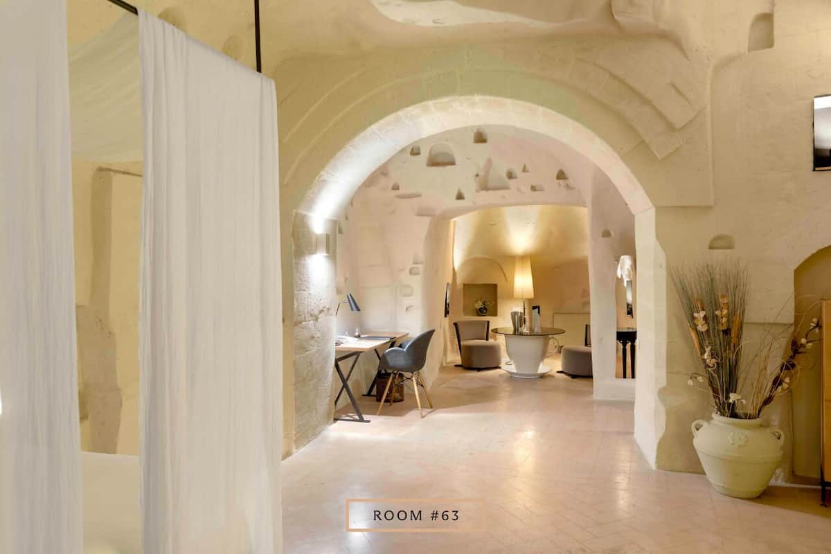A room with an arched doorway leading to another space, a dining area with a table and chairs, and a large vase with dried plants.