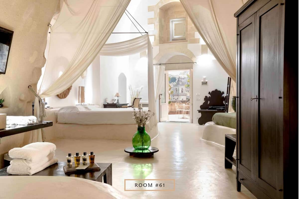 A spacious room with a bed, a vanity with toiletries, a green vase, and a view of a building through an archway.