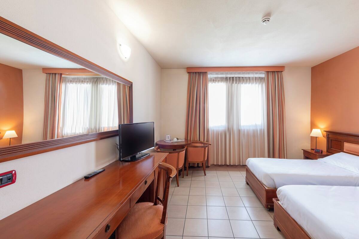 A hotel room with two beds, a desk with a TV, a chair, a small table, and a lamp.