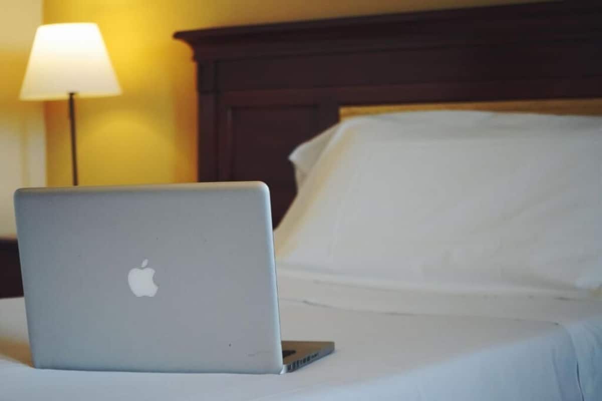 A laptop on a bed with a wooden headboard and a lamp in the background.