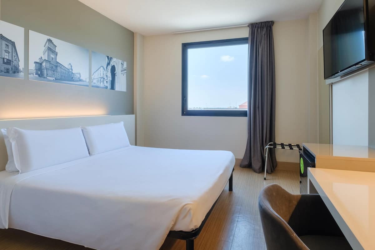 A hotel room with a bed, a desk, a TV, a window with curtains, and wall art.