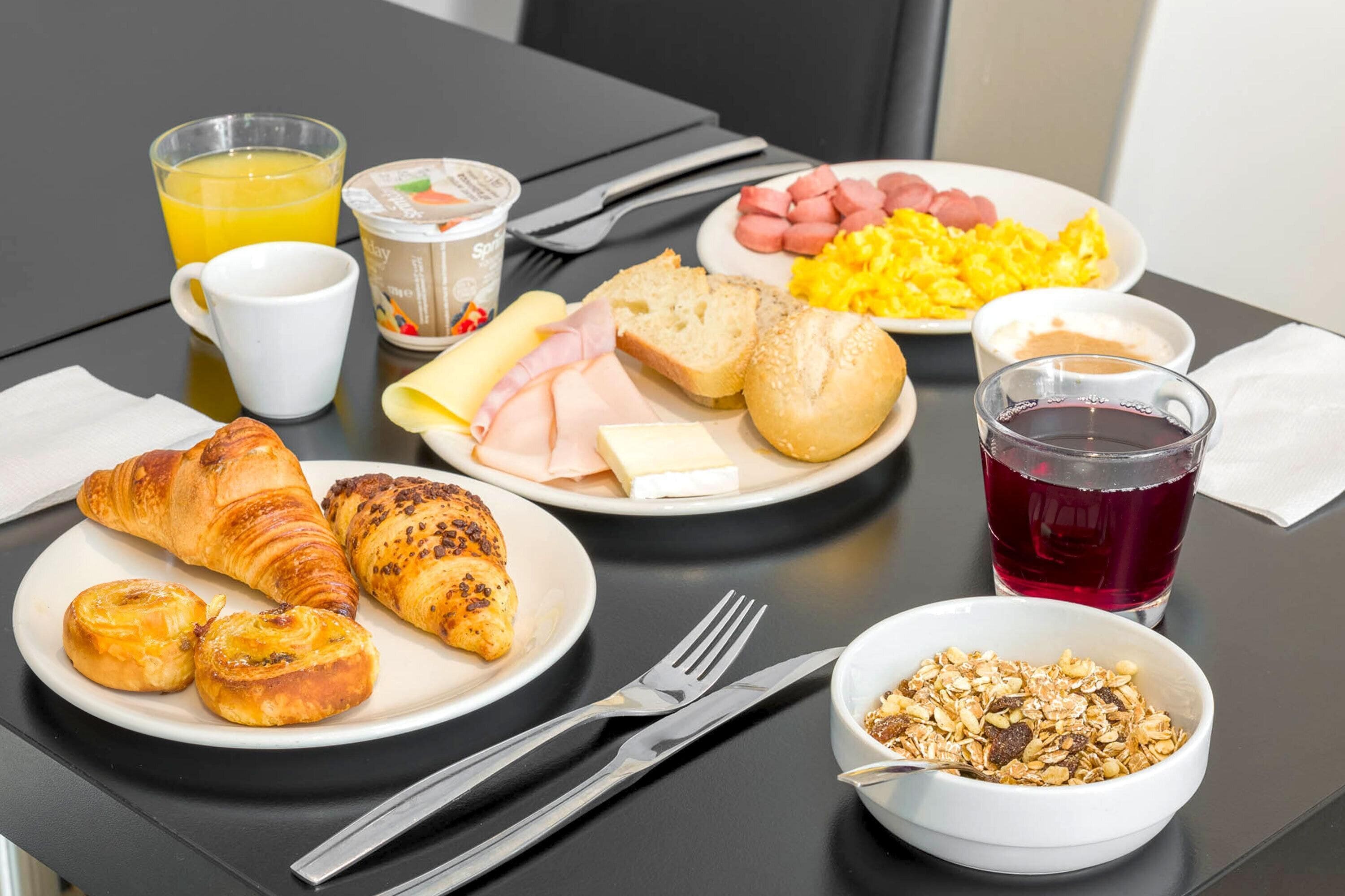 A breakfast table with croissants, ham, cheese, bread, scrambled eggs, sausages, orange juice, a glass of red juice, and a bowl of cereal.