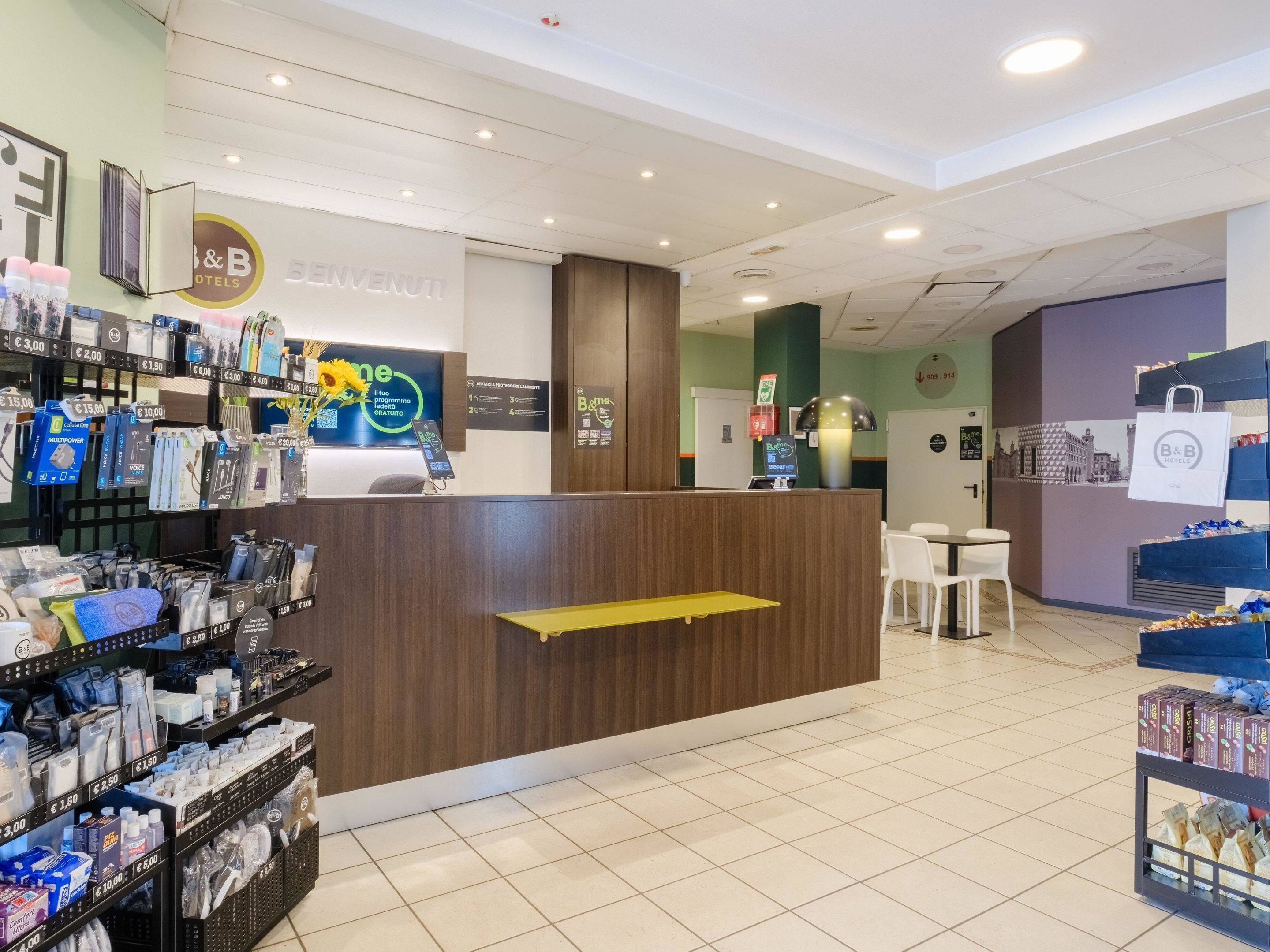 Un'area reception di un hotel con un bancone in legno, un chiosco self-service e diversi articoli in vendita sugli scaffali.