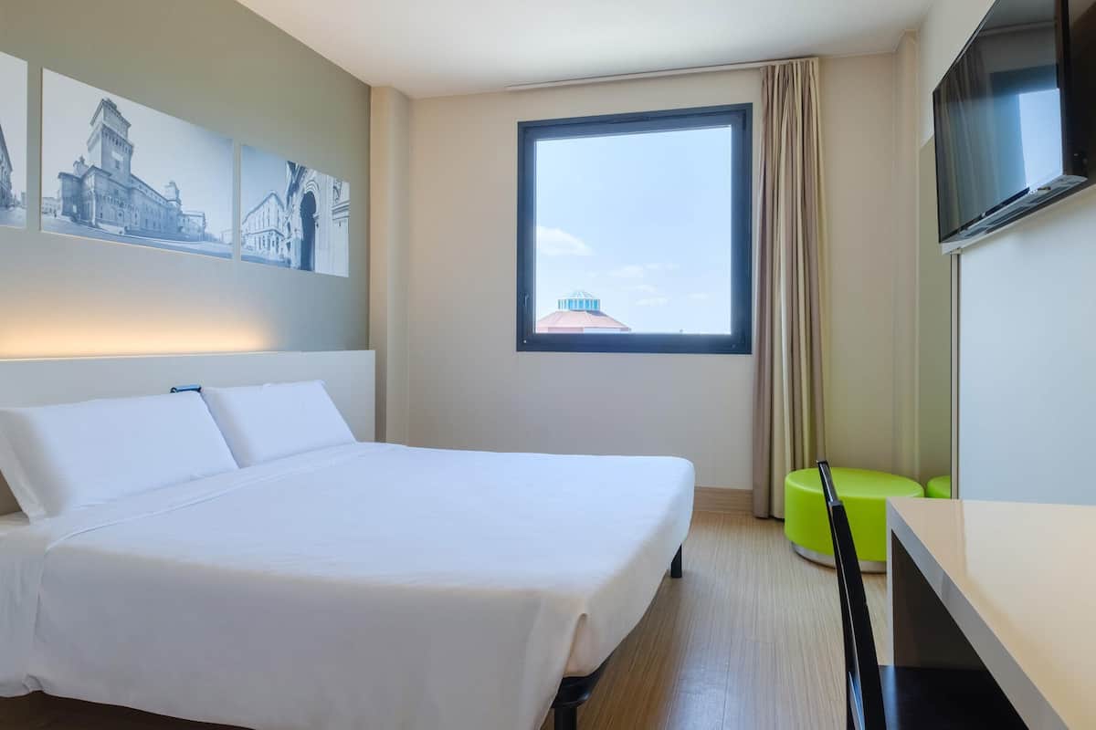 A hotel room with a bed, a TV, a window with a view, and a green chair.