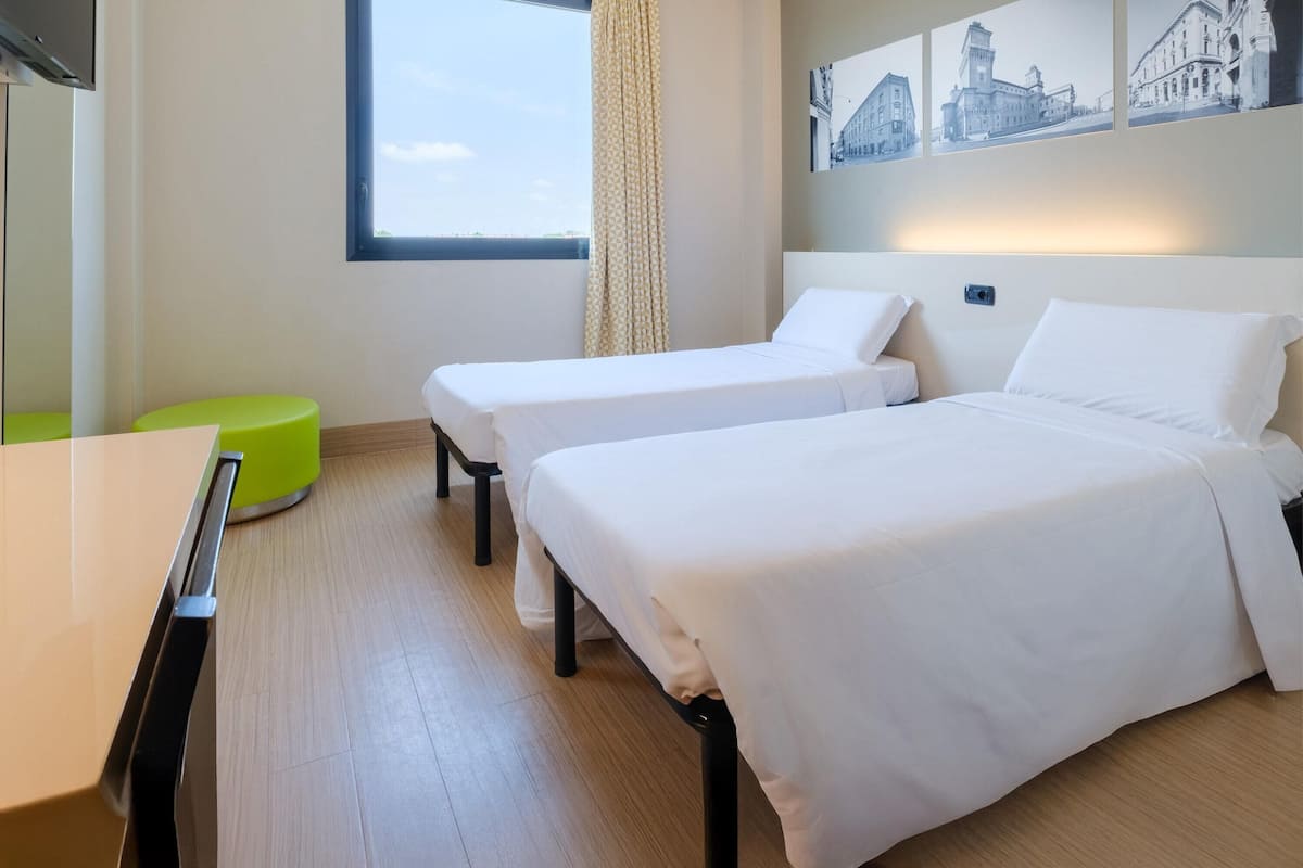 A hotel room with two single beds, a desk, a green stool, and a window with a view of buildings.