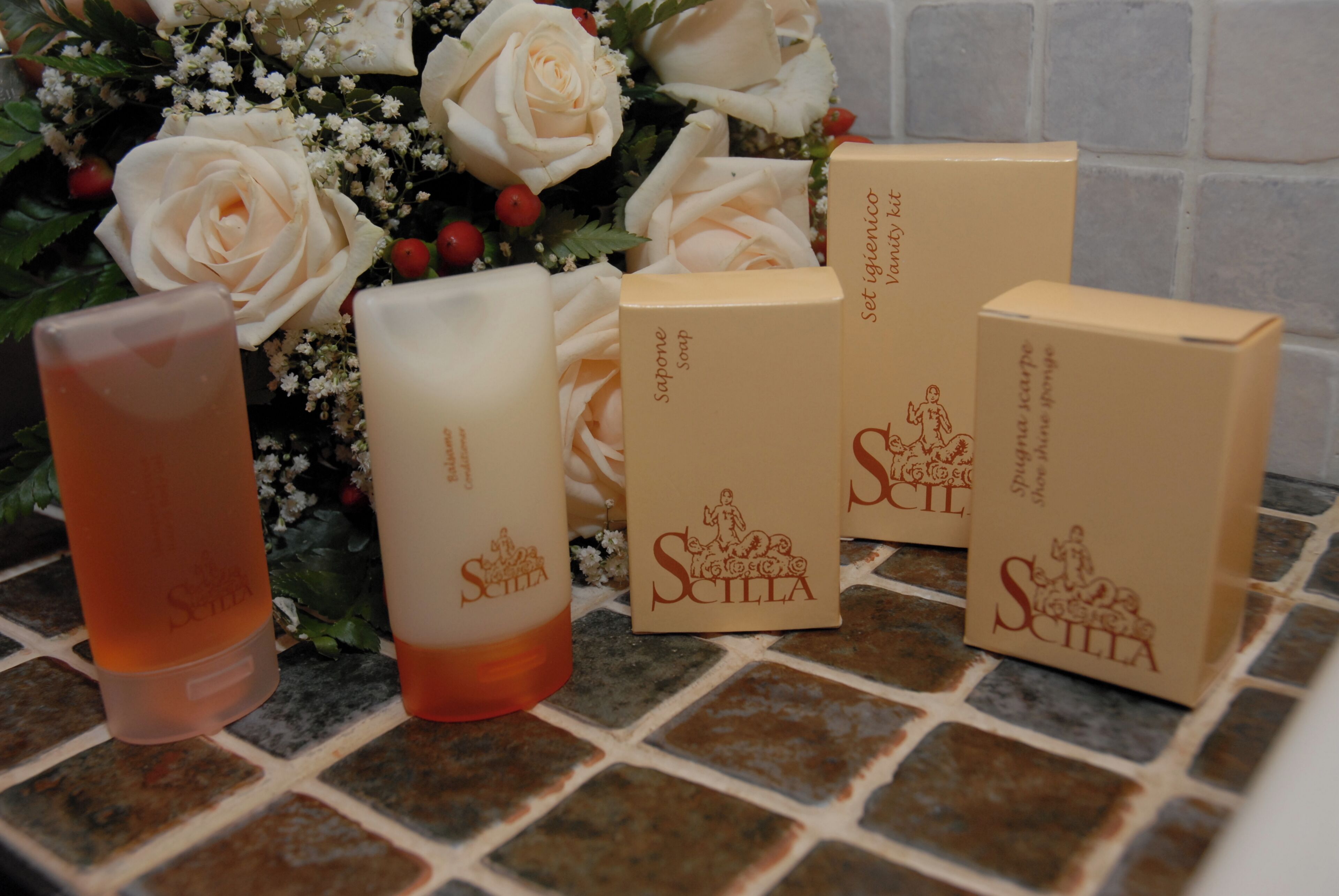A set of toiletries including a soap bar, a shampoo bottle, and a shower gel container, arranged on a tiled surface with a decorative arrangement of white roses in the background.