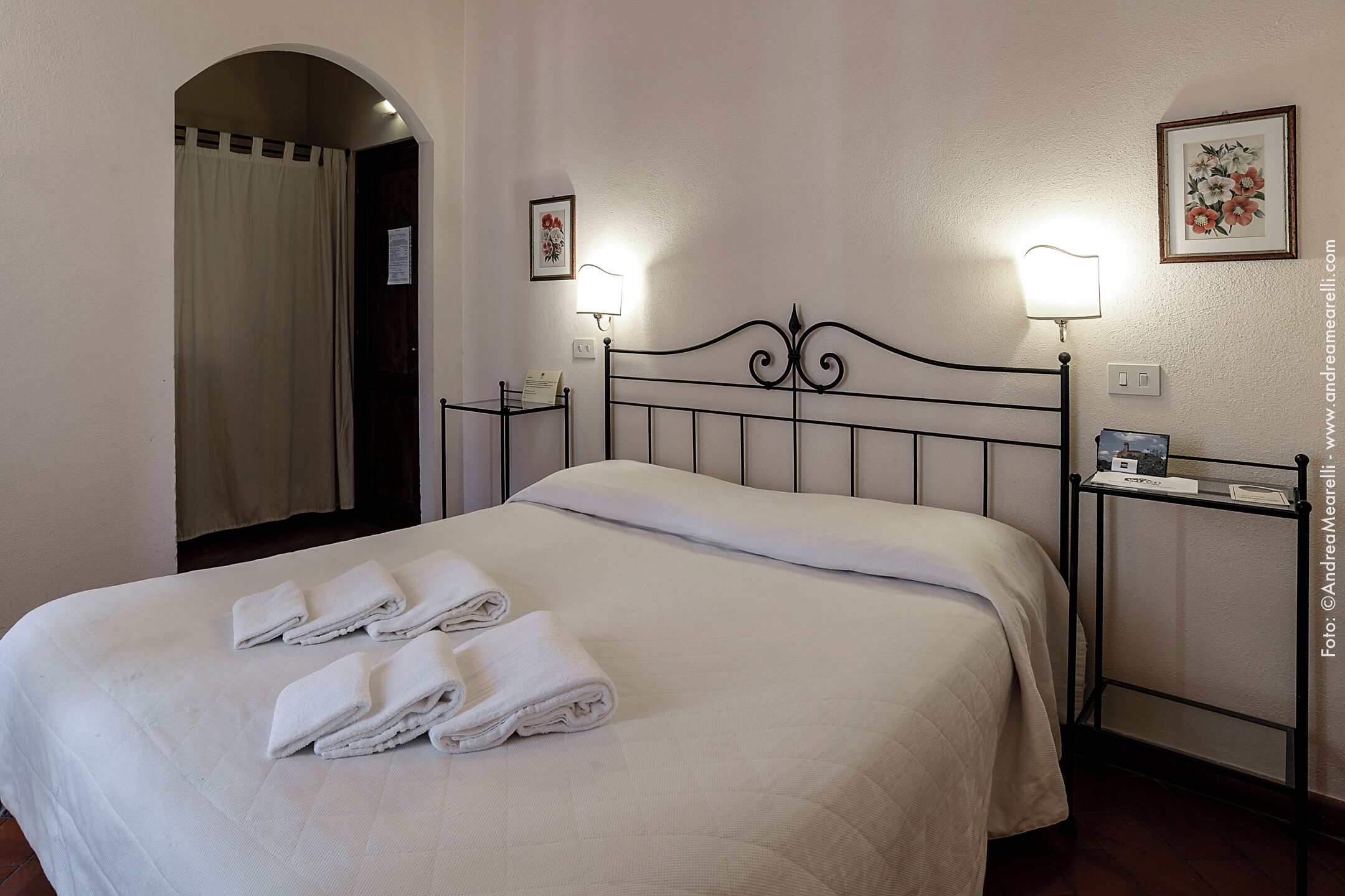 A neatly made bed with white linens and towels, a black metal headboard, two bedside tables with lamps, and a framed floral picture on the wall.