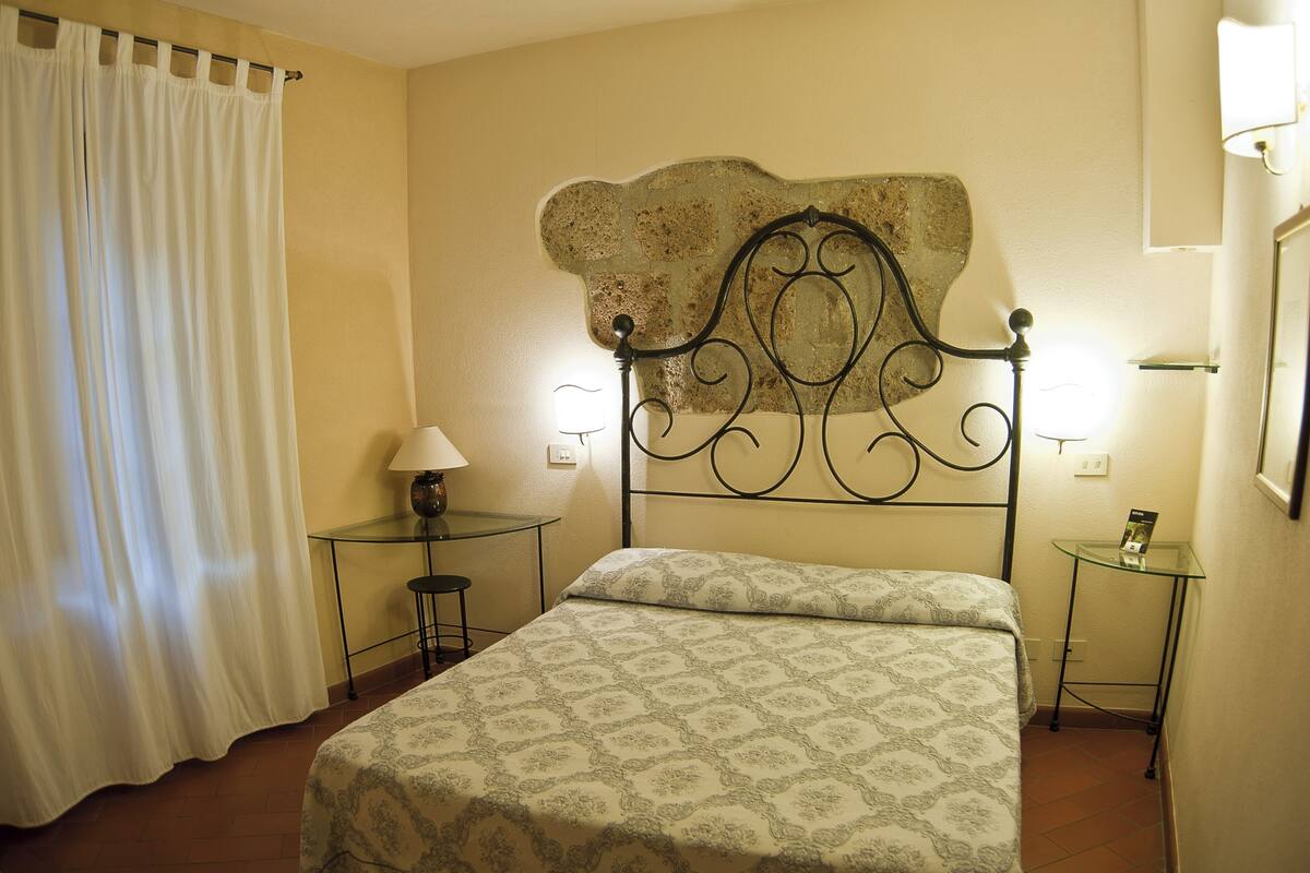 A bedroom with a bed, a nightstand, a lamp, a small table, and a curtain.