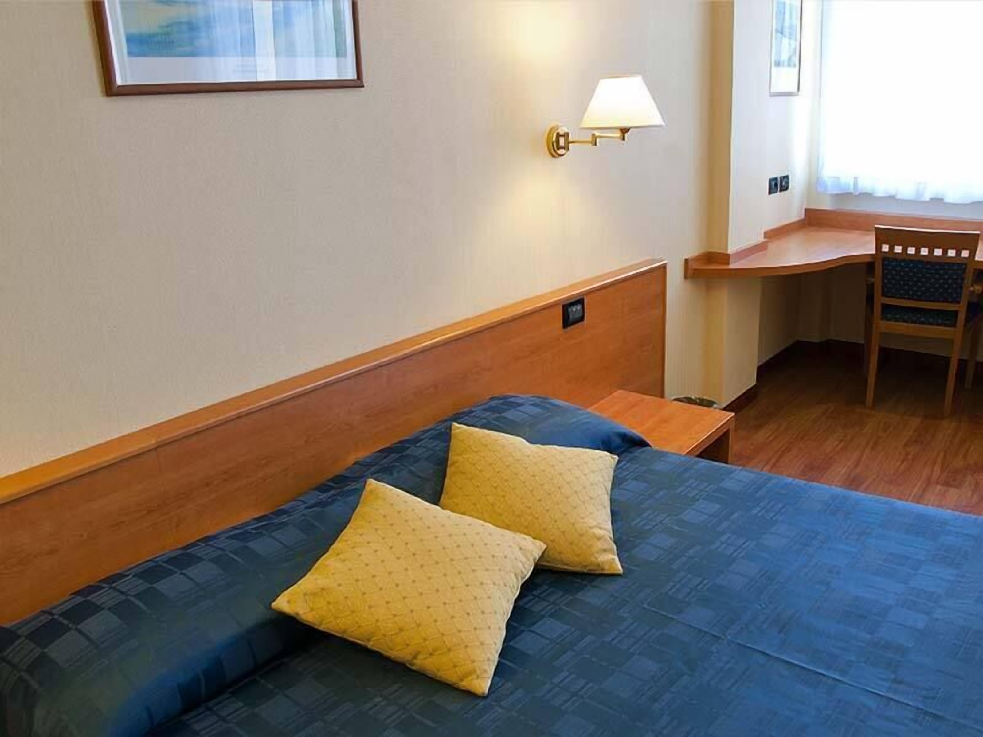 A hotel room with a bed, two pillows, a nightstand, a desk, and a chair.