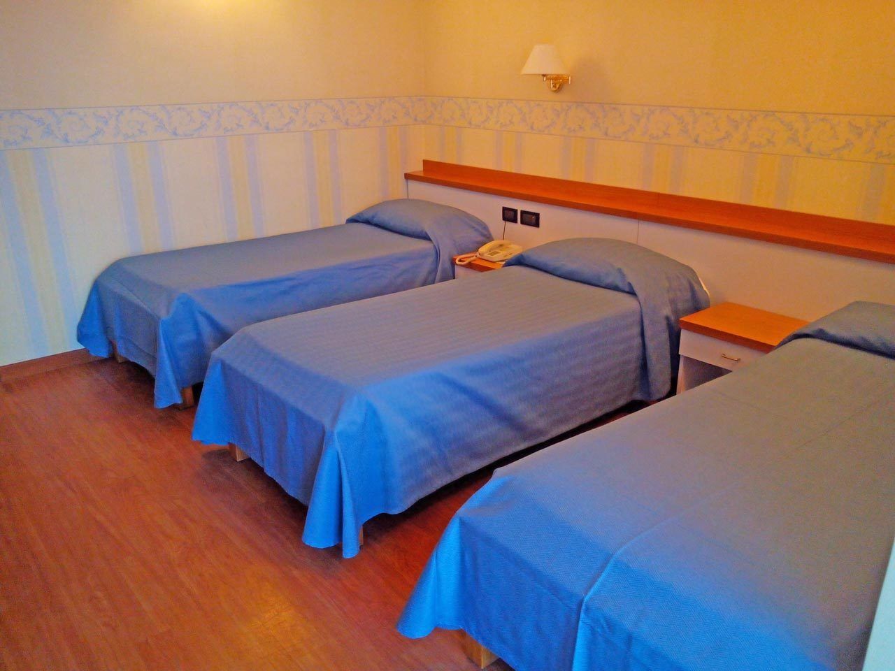 A room with three single beds, each with a blue bedspread and matching pillows. There is a wooden headboard, a bedside table with a lamp, and a telephone on the wall.