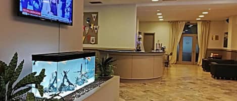 Un hall d’hôtel comprenant un comptoir d’accueil, un téléviseur fixé au mur et un grand aquarium avec des plantes aquatiques.