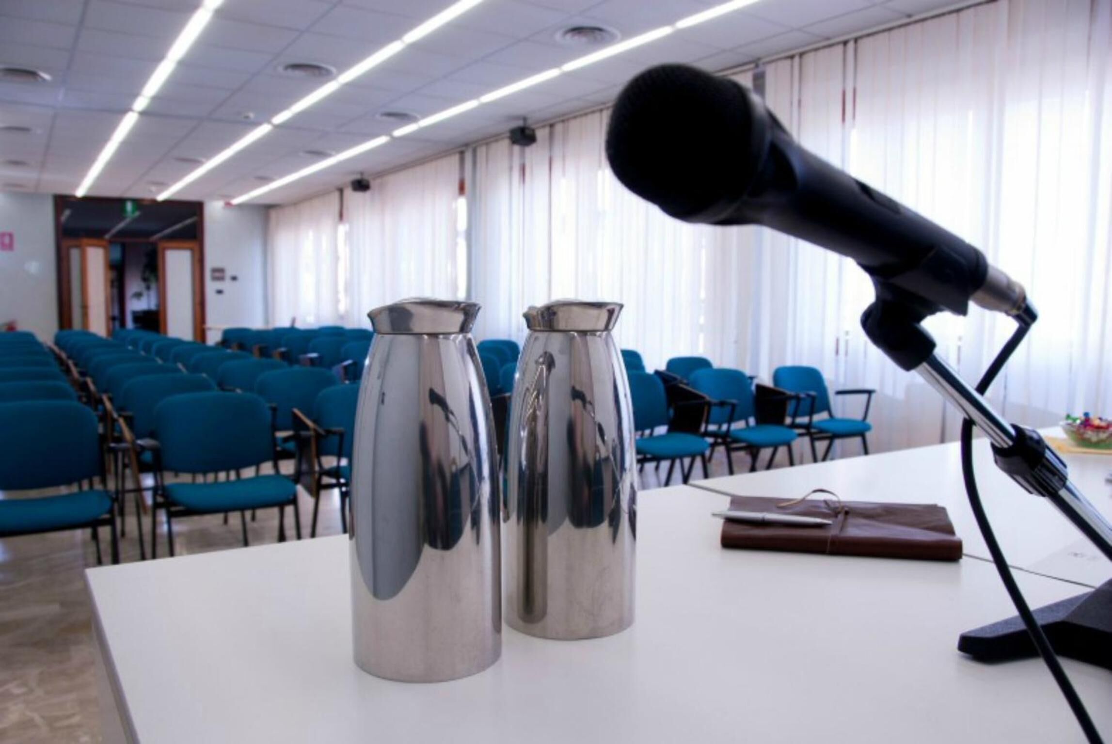 A conference room with rows of blue chairs, a white table with two metallic vases and a microphone, and a door in the background.