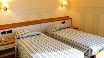 Une chambre d’hôtel avec deux lits simples, chacun doté de draps à rayures, d’une tête de lit en bois et d’une lampe de chevet. Une table de chevet en bois, sur laquelle se trouve un téléphone, est placée de chaque côté des lits.