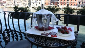 A table with a bottle, two glasses, a plate of strawberries, and a dessert on a balcony overlooking a canal with buildings in the background. - Palazzo Barbarigo Sul Canal Grande (Venice)