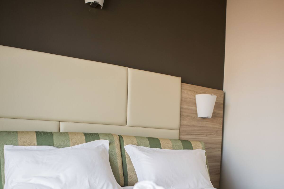 Un lit bien fait, avec du linge de lit blanc et une couvre-lit à motifs verts, une tête de lit en bois clair et un luminaire fixé au mur.