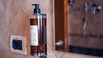 Una bottiglia di PRIJA detergente corpo su un ripiano del bagno con uno schienale piastrellato e una doccia sullo sfondo.