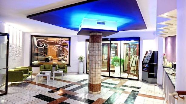 A lobby with a tiled floor, green chairs, a white coffee table, a pillar, and a glass door leading outside. - Hotel Prestige (Montesilvano)