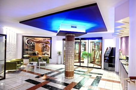A lobby with a tiled floor, green chairs, a white coffee table, a pillar, and a glass door leading outside.