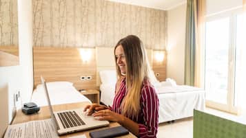 Habitación de hotel con cama, escritorio con computadora portátil, mesita de noche con lámpara y ventana con cortinas.