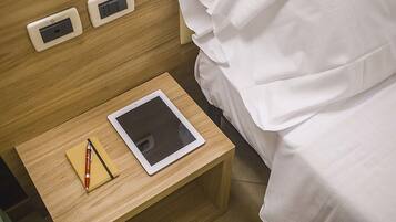 Una cama bien hecha con sábanas blancas, una mesita de noche de madera con una tableta y un bolígrafo, y dos enchufes en la pared.
