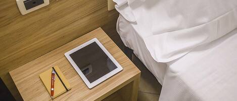 Un lit bien fait, avec du linge de lit blanc, une table de chevet en bois sur laquelle se trouvent une tablette et un stylo, et deux prises électriques sur le mur.