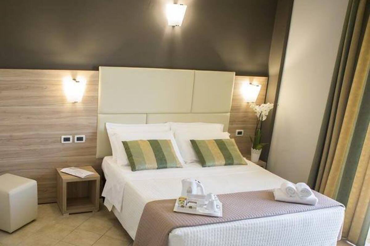 Une chambre d’hôtel avec un lit, des tables de chevet, une commode, une lampe de chevet, une fenêtre avec des rideaux et une petite plante en pot.