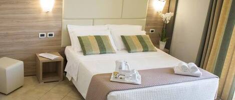 Quarto de hotel com cama, mesas de cabeceira, luminária, janela com cortinas e um pequeno vaso de planta.