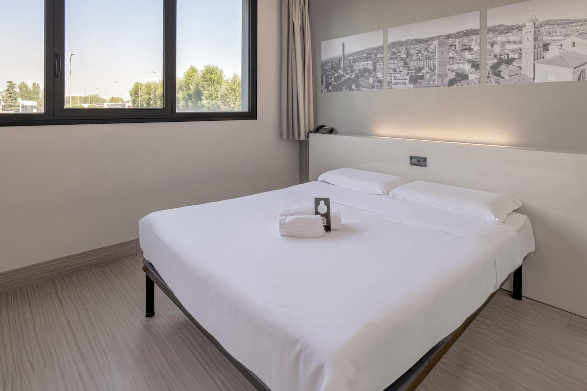A neatly made bed with white linens and towels, a cityscape view through a large window, and a wall-mounted light fixture.