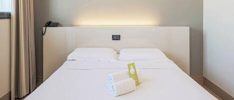 Habitación de hotel con una cama grande, ropa de cama blanca, vista panorámica de la ciudad, un teléfono en la mesita de noche y una etiqueta verde sobre la cama.