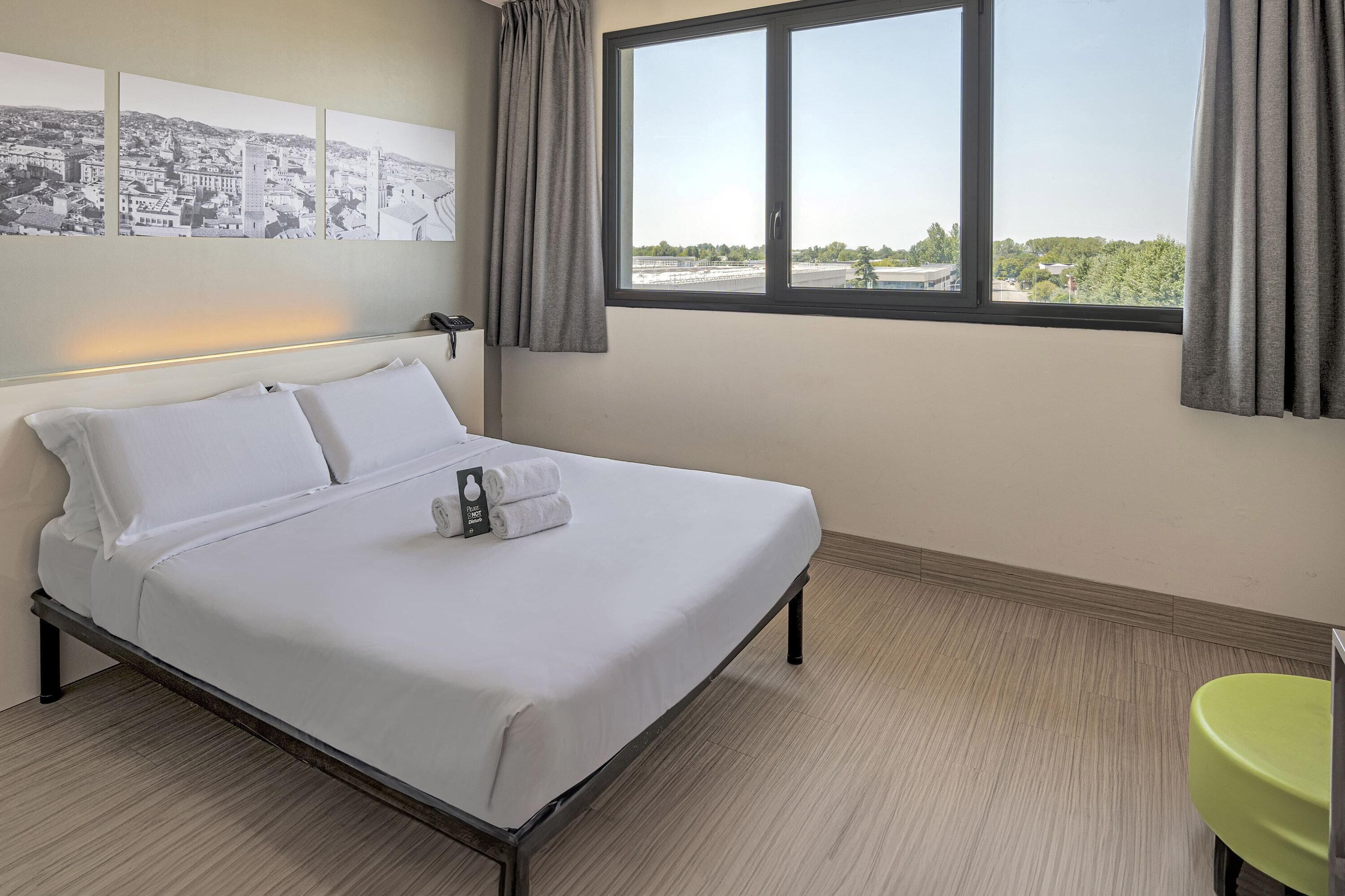 Habitación de hotel con cama, ventana con cortinas, una silla verde y una pared con tres fotografías en blanco y negro de paisajes urbanos.