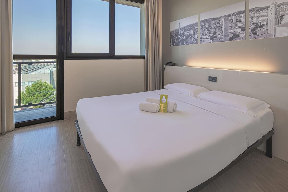 A hotel room with a large bed, a view through a window, and a cityscape mural on the wall.