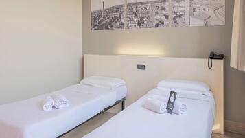A hotel room with two single beds, each with a white bedspread and rolled towels. There is a wall-mounted phone and a small nightstand with a lamp.