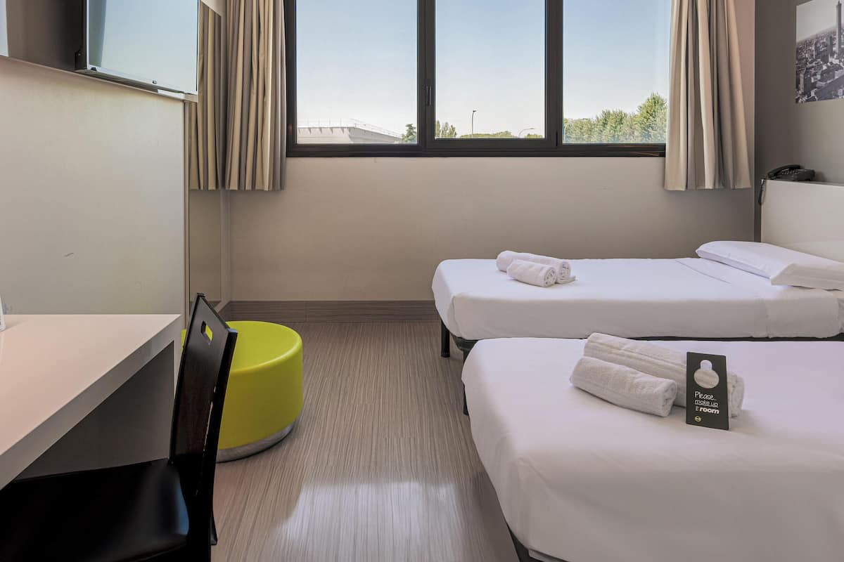 A hotel room with two beds, a desk, a chair, and a yellow ottoman.