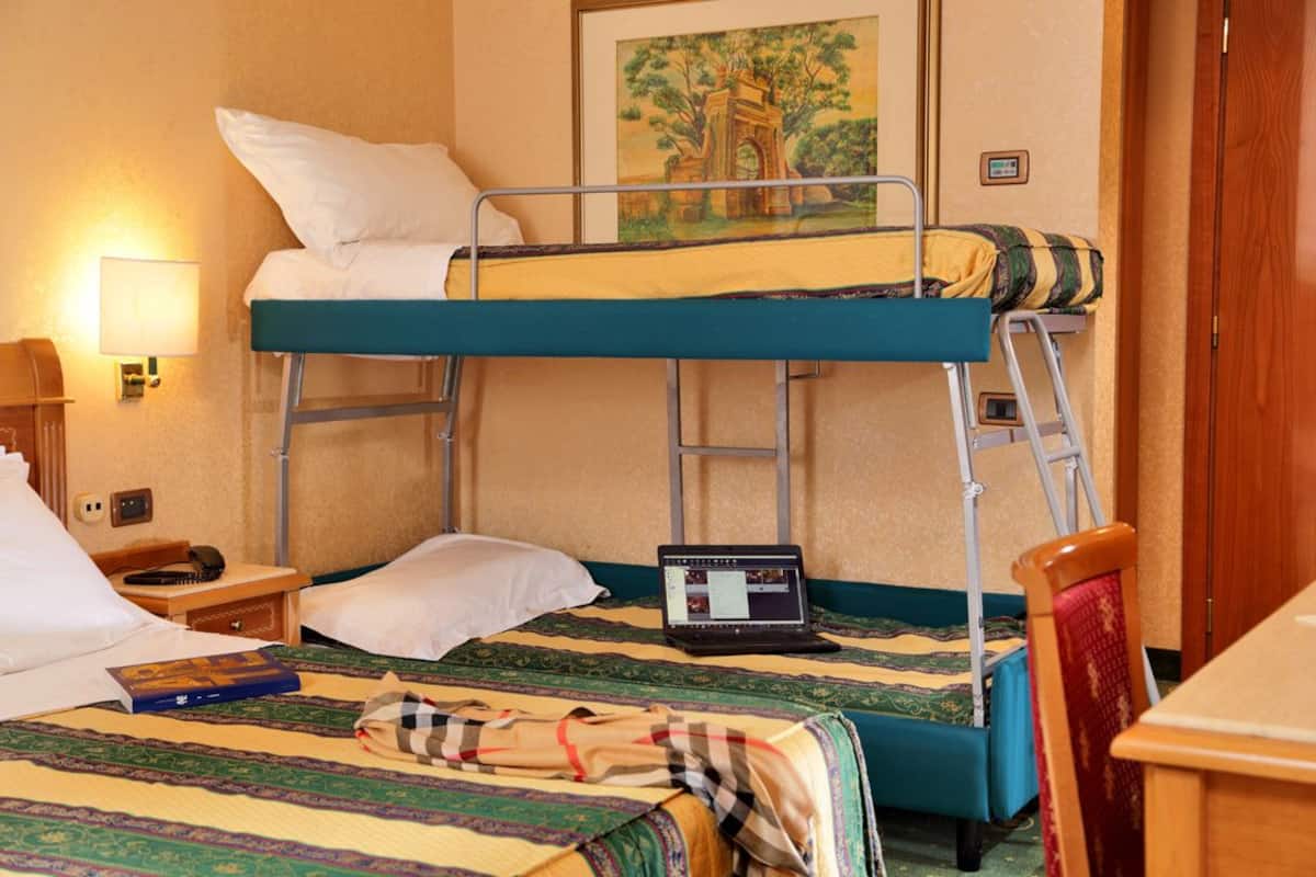 A bunk bed room with a laptop on the lower bunk, a bedside table with a lamp, and a painting on the wall.