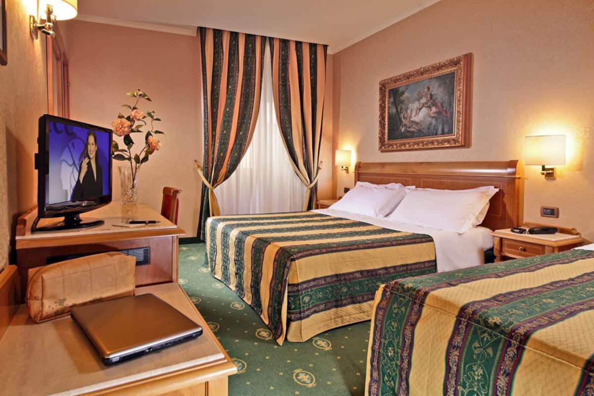 A hotel room with two beds, a desk with a laptop, a television, and a painting on the wall.