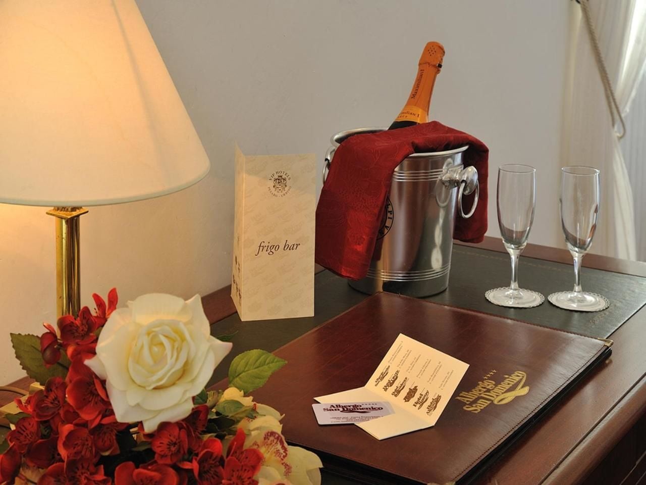 A hotel room with a bedside table featuring a lamp, a bouquet of flowers, a minibar, two glasses, a bottle of champagne, and a card.