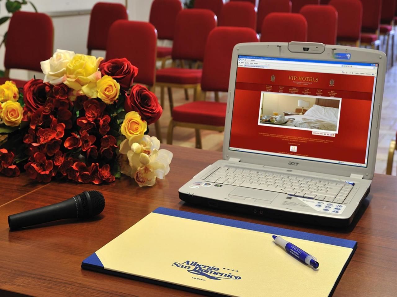A laptop on a wooden table displaying a hotel website, a microphone, a pen, and a bouquet of flowers.