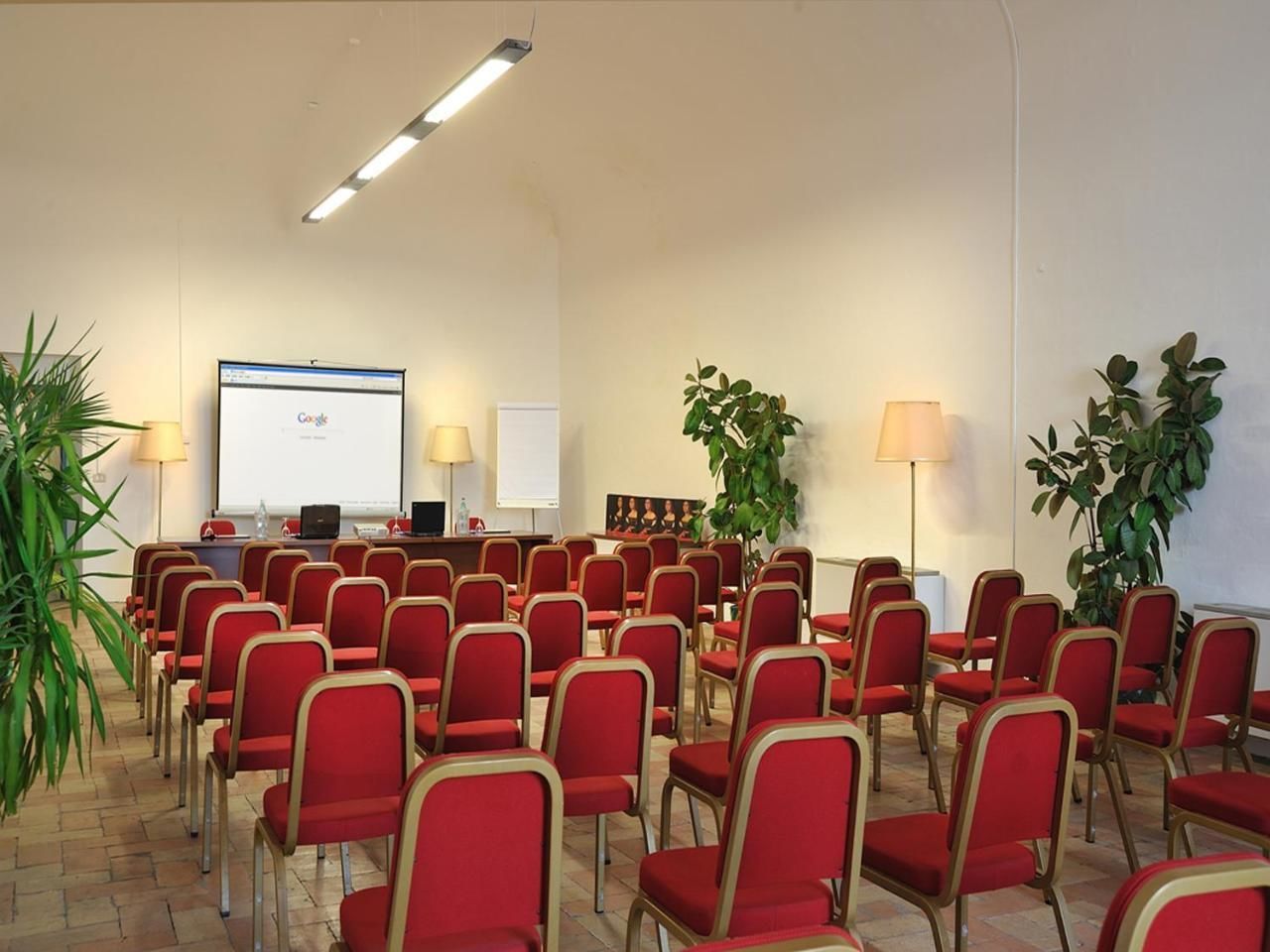 A conference room with rows of red chairs, a projector screen, and a laptop on a desk.