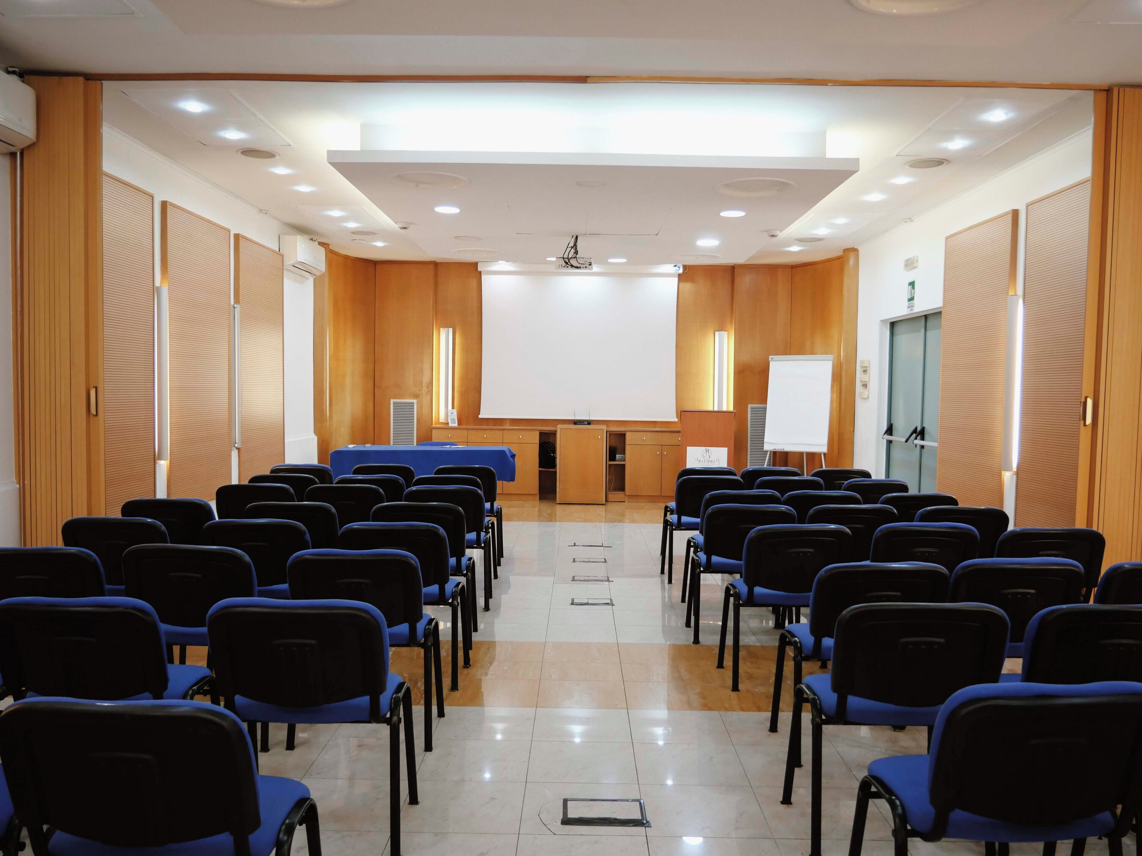 Konferenssihuone, jossa on riveissä sinisiä tuoleja, valkoinen projektorikangas ja puupanelointi.