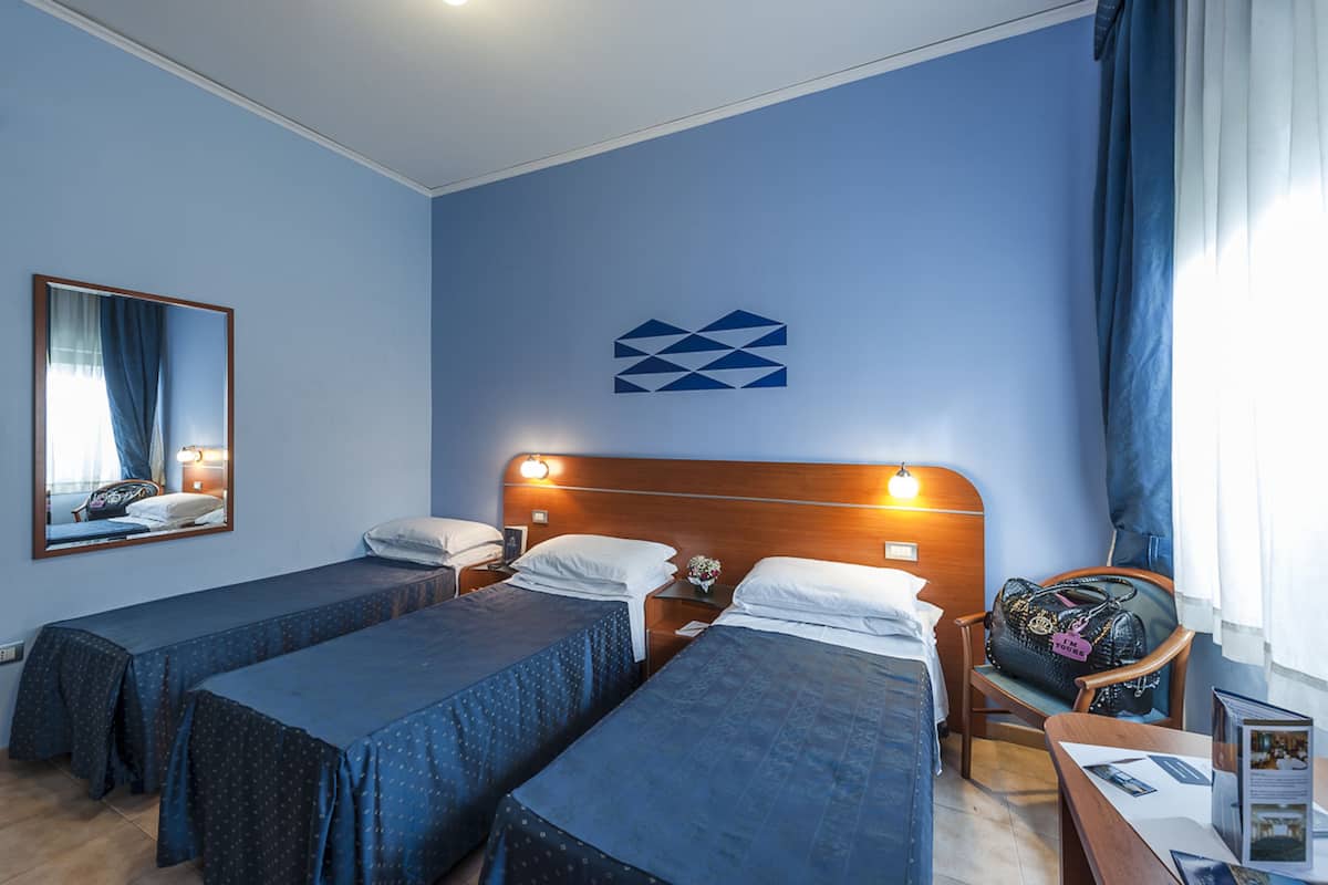 A hotel room with two single beds, a wooden headboard, a nightstand with a lamp, a chair, and a table with a book and a brochure.